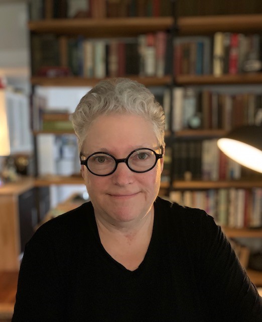 Michele Eodice has short, gray hair and glasses and is smiling. They are wearing a black shirt and are seated in a cozy room with book-filled shelves in the background. A warm lamp softly lights the space.